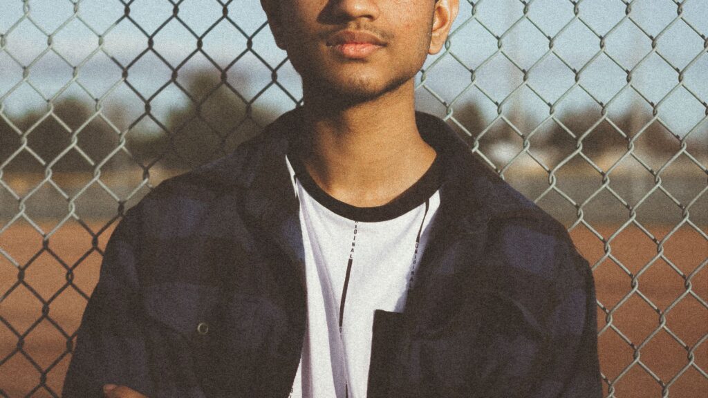 A teenage boy, head and shoulders, cropped below the eyes. He's wearing a black jacket and white t-shirt and stands in front of a metal fence
